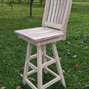 Puede incluir: Un taburete de bar de madera con un acabado de madera clara. El taburete tiene un respaldo de listones y un asiento cuadrado. El taburete está de pie sobre un césped.