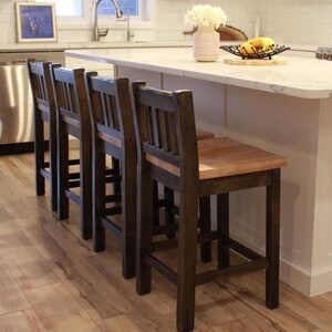 May include: Four wooden bar stools with dark brown frames and natural wood seats are arranged in a row. The stools have a slatted back design and are positioned in front of a white kitchen island with a light-coloured countertop.