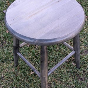 May include: A grey wooden stool with a round seat and four legs. The stool is made of solid wood and has a rustic finish.