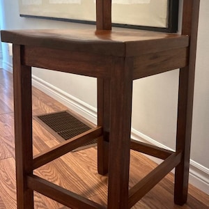 Puede incluir: Un taburete de bar de madera marrón oscuro con un diseño sencillo. El taburete tiene un asiento curvo y un respaldo recto.