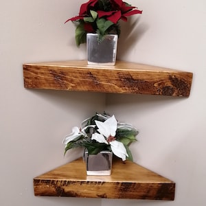 Puede incluir: Dos estantes de esquina de madera con un acabado desgastado. El estante superior tiene una planta de poinsettia roja en una maceta plateada. El estante inferior tiene una planta de poinsettia blanca en una maceta plateada.