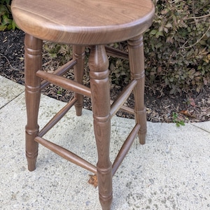 May include: A wooden stool with a round seat and four legs. The stool is made of a light brown wood and has a classic design. The legs are connected by horizontal supports for added stability. The stool is suitable for various settings.