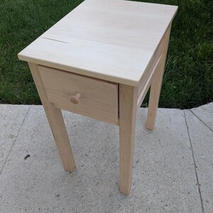 May include: A light-coloured wooden side table with a single drawer and a round knob. The table has four angled legs and a rectangular top. The table is approximately 61 cm tall.