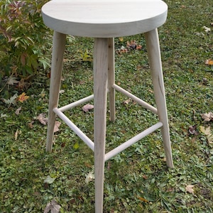 May include: A light-coloured wooden stool with a round seat and four angled legs. The stool has a simple, minimalist design and is made of unfinished wood. The legs are connected by horizontal supports near the base. The stool is approximately 61 cm tall.