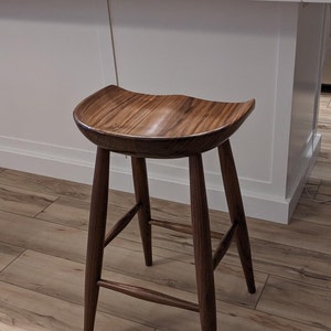 May include: A dark brown wooden bar stool with a curved seat and four legs. The stool is made of solid wood and has a natural finish.
