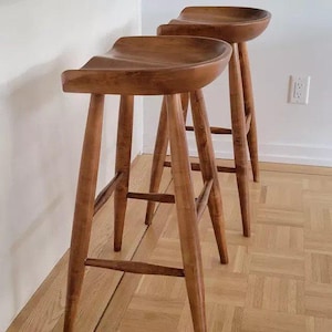 May include: Two tall bar stools crafted from medium-toned wood, each featuring a contoured, saddle-shaped seat and four tapered legs connected by horizontal support rungs. The stools have a smooth, polished finish highlighting the natural wood grain.