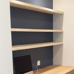 Puede incluir: Una unidad de estantería empotrada de madera clara y blanca con tres estantes contra una pared azul oscuro. Un monitor de computadora está sobre un escritorio de madera clara debajo de los estantes.