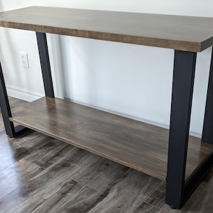 Puede incluir: Una mesa consola de madera marrón con un marco de metal negro y un estante inferior. La mesa tiene una tapa rectangular y está soportada por cuatro patas.
