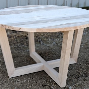 May include: A circular wooden table with a light-coloured, natural wood grain top. The table features a distinctive base design with angled legs that intersect in the centre. The table is made of solid wood and has a natural finish.