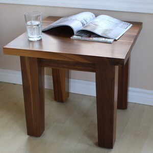 May include: A dark brown wooden end table with a simple, rectangular design. The table has four legs and a smooth, flat top. A glass of water and a magazine are sitting on the table.