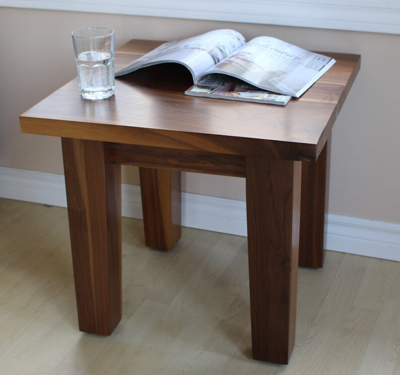 May include: A dark brown wooden end table with a simple, rectangular design. The table has four legs and a smooth, flat top. A glass of water and a magazine are sitting on the table.