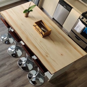 Könnte beinhalten: Eine helle Kücheninsel aus Holz mit einer Live-Edge-Arbeitsplatte und vier Barhockern. Die Insel hat einen weißen Sockel und eine kleine Holzkiste mit dem Text "Le Marché" darauf.