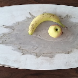 Puede incluir: Una bandeja de servir ovalada de madera blanca con un patrón de veta de madera natural. Una banana amarilla y una manzana roja se colocan en la bandeja.