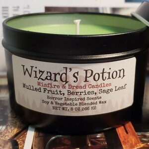 May include: A black metal candle tin with a green candle and a white wick. The label reads "Wizard's Potion" with scent notes. The lid is partially open. The candle is 8 oz.