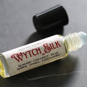 Puede incluir: Un frasco roll-on de vidrio transparente con una tapa negra. La etiqueta dice "WYTCH SILK" en escritura roja, con notas de fragancia enumeradas a continuaci&oacute;n. El frasco contiene un l&iacute;quido amarillo claro.