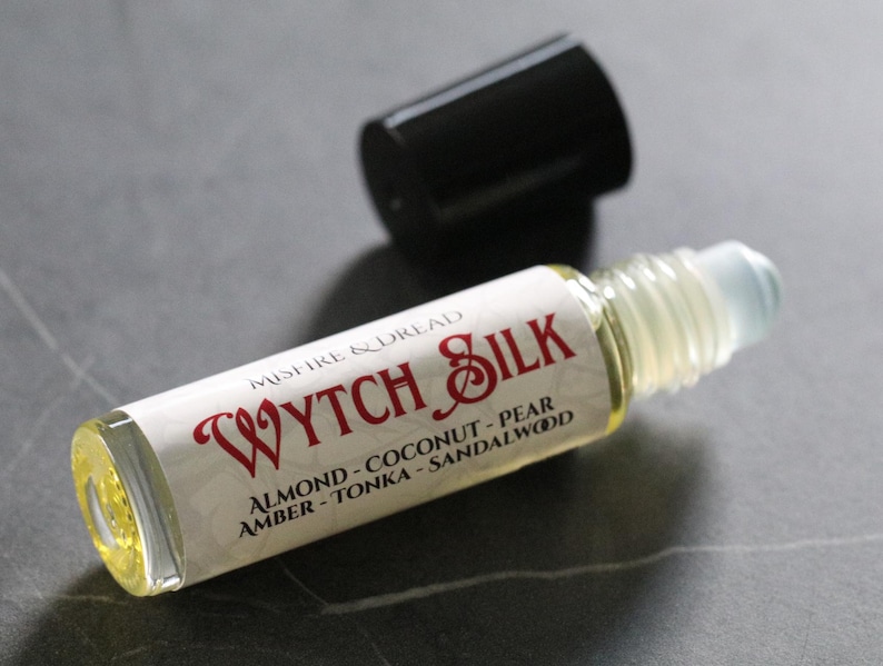 Puede incluir: Un frasco roll-on de vidrio transparente con una tapa negra. La etiqueta dice "WYTCH SILK" en escritura roja, con notas de fragancia enumeradas a continuaci&oacute;n. El frasco contiene un l&iacute;quido amarillo claro.