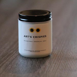 May include: A white candle in a glass jar with a black lid. The label features a yellow sunglasses icon, the text ART'S CRISPIES, and the words RICE CRISPY TREATS - MARSHMALLOW - VANILLA. Additional text includes Soy & Vegetable Wax.