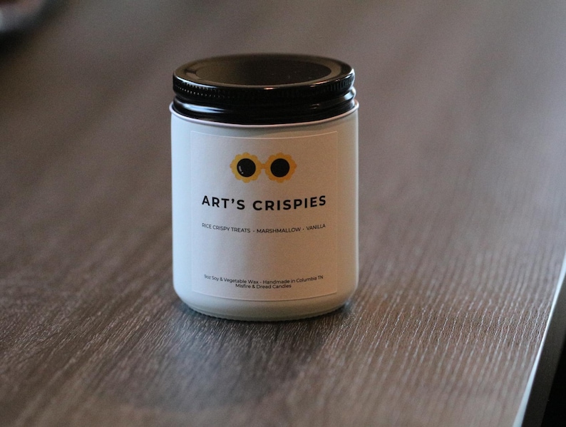 May include: A white candle in a glass jar with a black lid. The label features a yellow sunglasses icon, the text ART'S CRISPIES, and the words RICE CRISPY TREATS - MARSHMALLOW - VANILLA. Additional text includes Soy & Vegetable Wax.