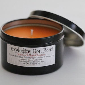 May include: A black metal tin candle with an orange wax interior and a white wick. The label reads "Exploding Bon Bons Minfire & Bread Candles Blood Orange, Pineapple, Cherry, Vanilla Horror Inspired Scents Soy & Vegetable Blended Wax NET WT. 8 OZ (226 g)"