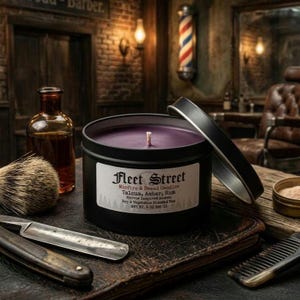 May include: A black candle tin with a purple wax interior and a white label that reads "Fleet Street". The candle is next to a straight razor, shaving brush, and a bottle of amber liquid. The candle is 8 oz.