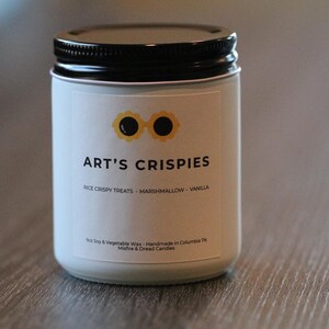 May include: A white candle in a glass jar with a black lid. The label features a yellow graphic of sunglasses above the text ART'S CRISPIES, followed by RICE CRISPY TREATS, MARSHMALLOW, VANILLA, and smaller text including Soy & Vegetable Wax.