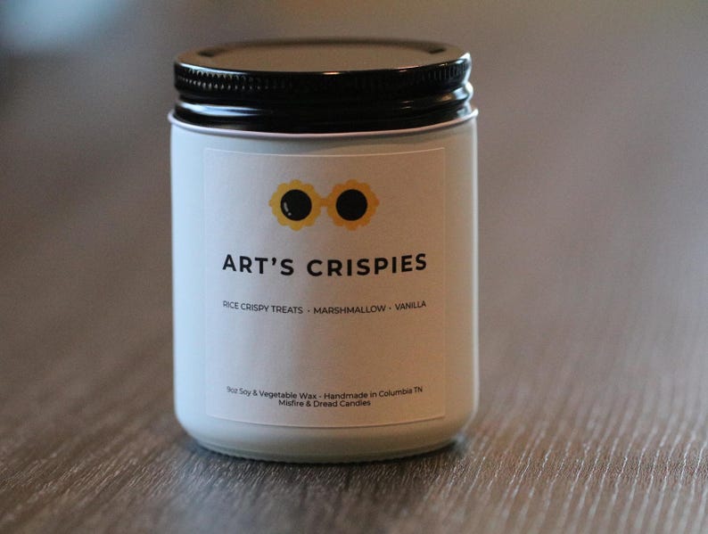 May include: A white candle in a glass jar with a black lid. The label features a yellow graphic of sunglasses above the text ART'S CRISPIES, followed by RICE CRISPY TREATS, MARSHMALLOW, VANILLA, and smaller text including Soy & Vegetable Wax.