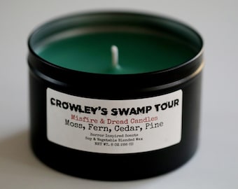 May include: A black candle tin with a green candle inside. The label reads "CROWLEY'S SWAMP TOUR" and lists scents like Moss, Fern, Cedar, and Pine. The candle is 8 oz (226 g) and made with soy and vegetable wax.