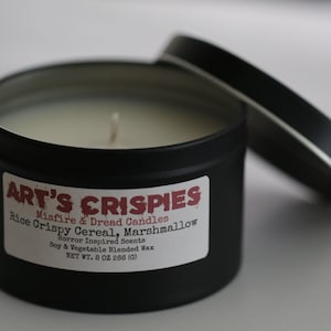 May include: A black metal tin candle with a white label reading ART'S CRISPIES, Misfire & Dread Candles, Rice Crispy Cereal, Marshmallow, Horror Inspired Scents, Soy & Vegetable Blended Wax, NET WT. 8 OZ 266 (G). The candle has a single wick and a lid.
