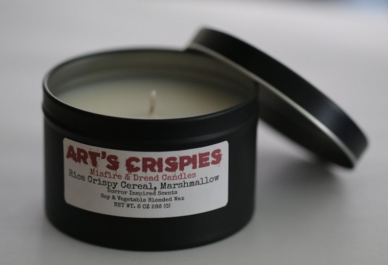 May include: A black metal tin candle with a white label reading ART'S CRISPIES, Misfire & Dread Candles, Rice Crispy Cereal, Marshmallow, Horror Inspired Scents, Soy & Vegetable Blended Wax, NET WT. 8 OZ 266 (G). The candle has a single wick and a lid.