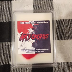 May include: A rectangular plastic clamshell container holding white wax melts. The label features the text Rice Crispy Treats, Marshmallow, and ARTS CRISPIES, with a split white and black background, red paint splatters, and two dark spheres.