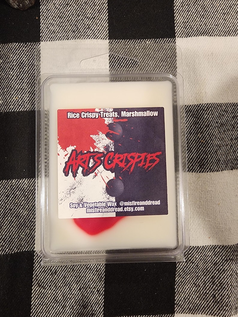 May include: A rectangular plastic clamshell container holding white wax melts. The label features the text Rice Crispy Treats, Marshmallow, and ARTS CRISPIES, with a split white and black background, red paint splatters, and two dark spheres.