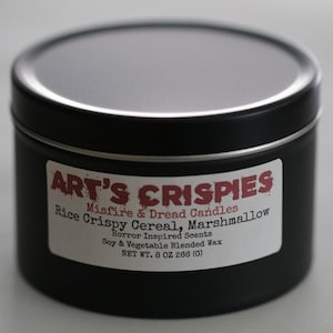 May include: A black cylindrical metal candle tin with a white label featuring the text ART'S CRISPIES, Misfire & Dread Candles, Rice Crispy Cereal, Marshmallow, Horror Inspired Scents, Soy & Vegetable Blended Wax, and NET WT. 8 OZ 266 (G).