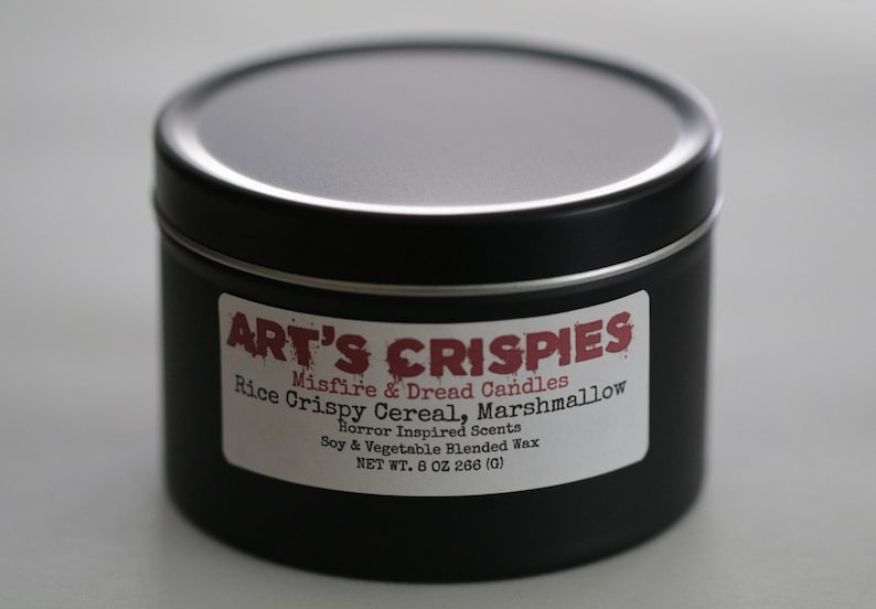 May include: A black cylindrical metal candle tin with a white label featuring the text ART'S CRISPIES, Misfire & Dread Candles, Rice Crispy Cereal, Marshmallow, Horror Inspired Scents, Soy & Vegetable Blended Wax, and NET WT. 8 OZ 266 (G).