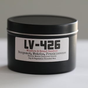 May include: A black candle tin with a white label that reads "LV-426 Misfire & Dread Candles" with scent descriptions. The candle is made with soy and vegetable blended wax. The tin is round with a matching black lid.