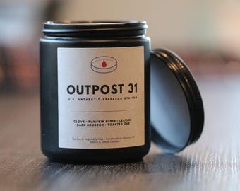 May include: A matte black candle jar with a white label featuring the text OUTPOST 31 U.S. ANTARCTIC RESEARCH STATION, a petri dish icon with a red drop, and scent notes of clove, pumpkin puree, leather, dark bourbon, and toasted oak.