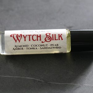 Puede incluir: Una peque&ntilde;a botella rectangular de vidrio de perfume "Wytch Silk" con tapa negra. La etiqueta muestra "MISTIRE & DREAD" sobre "WYTCH SILK" en escritura roja. Las notas de fragancia incluyen almendra, coco, pera, &aacute;mbar, haba tonka y s&aacute;ndalo.