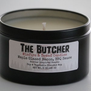 May include: A black metal tin candle with a white label that reads "THE BUTCHER Misfire & Dread Candles Maple Glazed Bacon, BBQ Sauce Horror Inspired Scents Soy & Vegetable Blended Wax NET WT. 8 OZ 266 (G)"