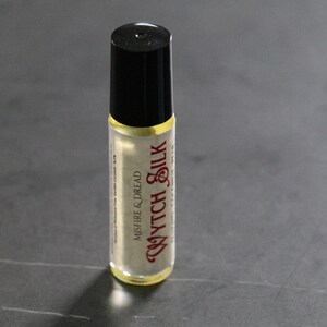 Puede incluir: Peque&ntilde;o frasco roll-on de vidrio transparente con tapa negra. El frasco contiene un l&iacute;quido de color claro y presenta el texto "Wytch Silk" en una fuente decorativa de color burdeos. Las palabras "Misfire & Dread" tambi&eacute;n est&aacute;n impresas.