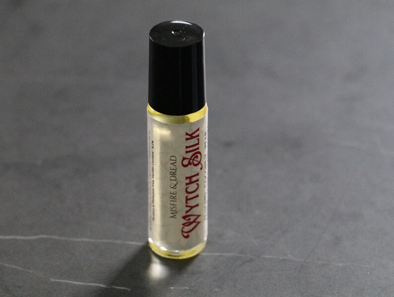 Puede incluir: Peque&ntilde;o frasco roll-on de vidrio transparente con tapa negra. El frasco contiene un l&iacute;quido de color claro y presenta el texto "Wytch Silk" en una fuente decorativa de color burdeos. Las palabras "Misfire & Dread" tambi&eacute;n est&aacute;n impresas.