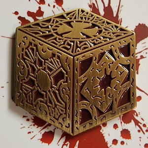 May include: A gold-colored metal cube with intricate designs and a red interior. The cube has a star-shaped design on one side. The cube is on a white background with red splatters.