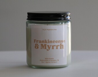 May include: A glass candle jar with a black lid. The candle is white and has a label that reads "Frankincense & Myrrh". The label also states "Joy & Vegetable Wax" and "M&D Candles Handcrafted in Columbia, TN. Burn Approx 40 Hours."