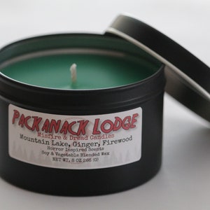 May include: A black metal tin candle with a green wax and a white wick. The label reads "Packanack Lodge Misfire & Dread Candles Mountain Lake, Ginger, Firewood Horror Inspired Scents Soy & Vegetable Blended Wax NET WT. 8 OZ 266 (0)"