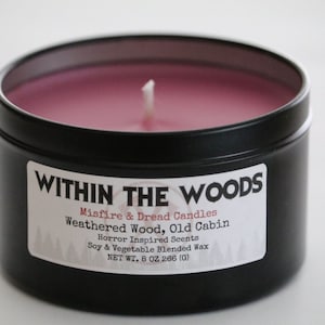 May include: A black metal tin candle with a pink wax candle inside. The label reads "WITHIN THE WOODS", "Misfire & Dread Candles", "Weathered Wood, Old Cabin", "Horror Inspired Scents", "Soy & Vegetable Blended Wax", and "NET WT. 8 OZ 266 (G)".