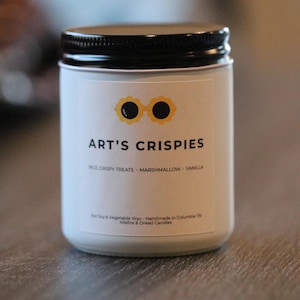 May include: A white candle in a glass jar with a black screw-top lid. The label features a yellow flower-shaped icon with black centres, the text ART'S CRISPIES, and the words RICE CRISPY TREATS - MARSHMALLOW - VANILLA printed in black.