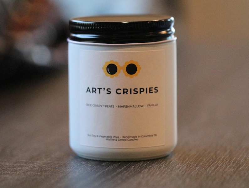 May include: A white candle in a glass jar with a black screw-top lid. The label features a yellow flower-shaped icon with black centres, the text ART'S CRISPIES, and the words RICE CRISPY TREATS - MARSHMALLOW - VANILLA printed in black.