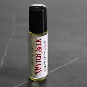 Puede incluir: Un frasco de perfume roll-on con tapa negra y cuerpo de vidrio transparente. La etiqueta dice "Wytch Silk" en una fuente roja estilizada, con "Misfire & Dread" arriba. Los ingredientes son almendras, coco, pera, &aacute;mbar, haba tonka y s&aacute;ndalo.