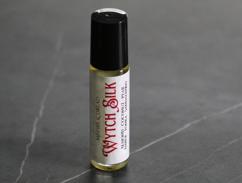 Puede incluir: Un frasco de perfume roll-on con tapa negra y cuerpo de vidrio transparente. La etiqueta dice "Wytch Silk" en una fuente roja estilizada, con "Misfire & Dread" arriba. Los ingredientes son almendras, coco, pera, &aacute;mbar, haba tonka y s&aacute;ndalo.