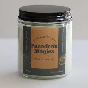 May include: A clear glass jar candle with a black lid. The label reads "Panadería Mágica" in brown and white text, with "baked to perfection" below. The candle is filled with white wax and is 8 oz.
