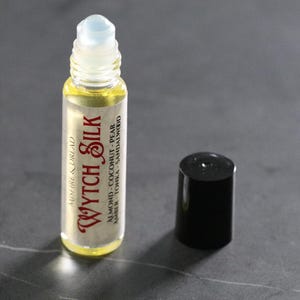 Puede incluir: Un frasco roll-on de vidrio transparente con una tapa de bola blanca y una tapa negra. El frasco contiene un l&iacute;quido amarillo y tiene las palabras "Wytch Silk" en una fuente decorativa. La etiqueta tambi&eacute;n enumera los ingredientes: almendras, coco, pera, &aacute;mbar, tonka y s&aacute;ndalo.