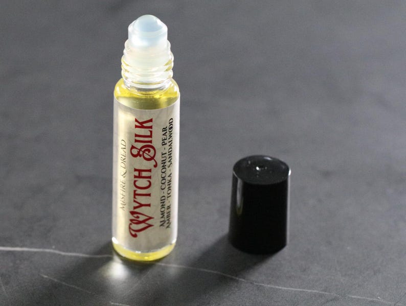 Puede incluir: Un frasco roll-on de vidrio transparente con una tapa de bola blanca y una tapa negra. El frasco contiene un l&iacute;quido amarillo y tiene las palabras "Wytch Silk" en una fuente decorativa. La etiqueta tambi&eacute;n enumera los ingredientes: almendras, coco, pera, &aacute;mbar, tonka y s&aacute;ndalo.